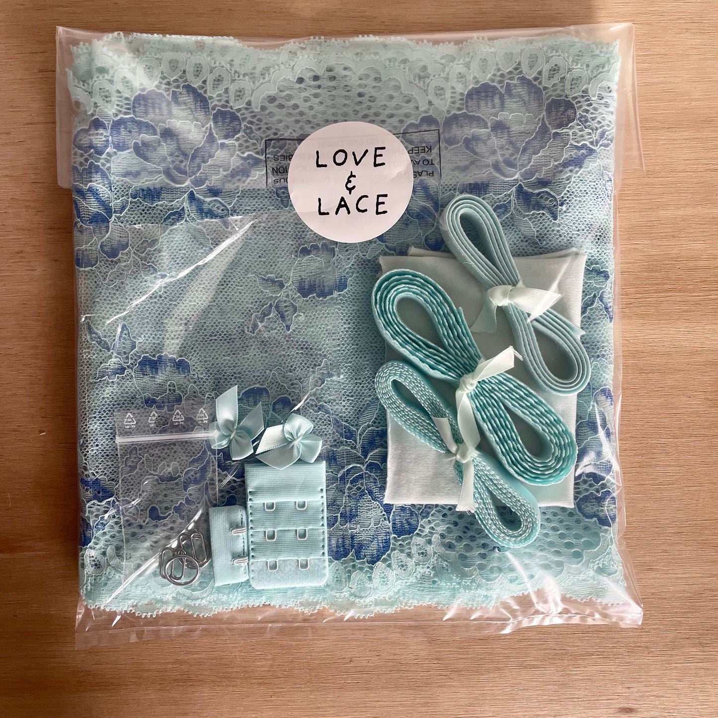 A packaged sewing or craft kit containing a light blue lace fabric, matching elastic and trims for bra making, all sealed in a clear plastic bag with a circular 'LOVE & LACE' label on a wooden surface.